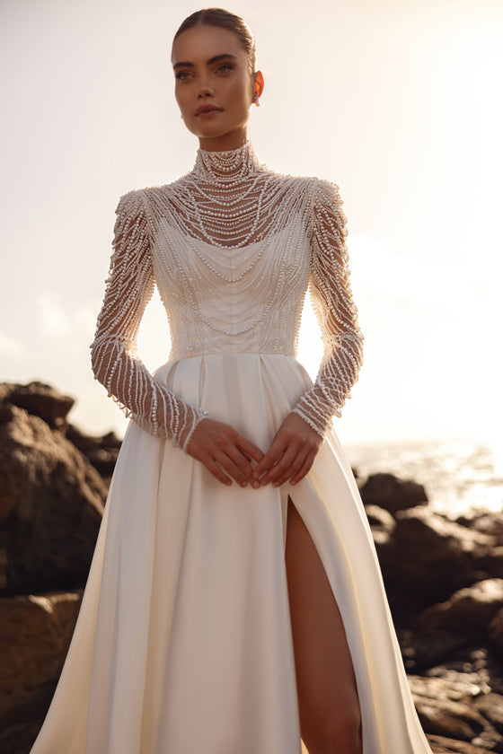 La blusa da sposa Alya, con lunghe maniche decorate con perle, abbinata a una gonna con spacco alto, esprime un look contemporaneo e prezioso.