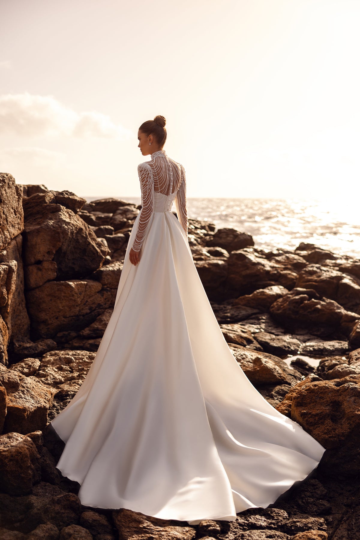La blusa da sposa Alya, con lunghe maniche impreziosite di perle e uno strascico scenografico, crea un look regale.