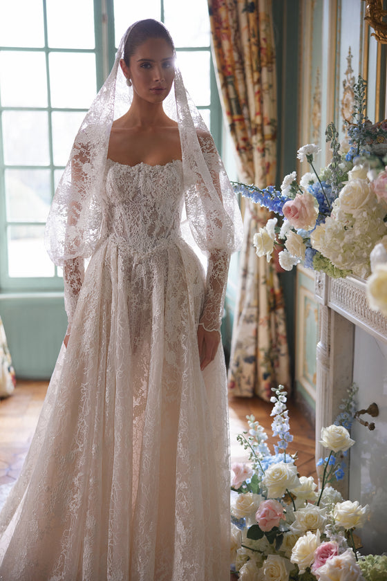 Abito da sposa Thetis – silhouette a trapezio con un corsetto trasparente, spalline in pizzo e una ricca gonna in pizzo.
