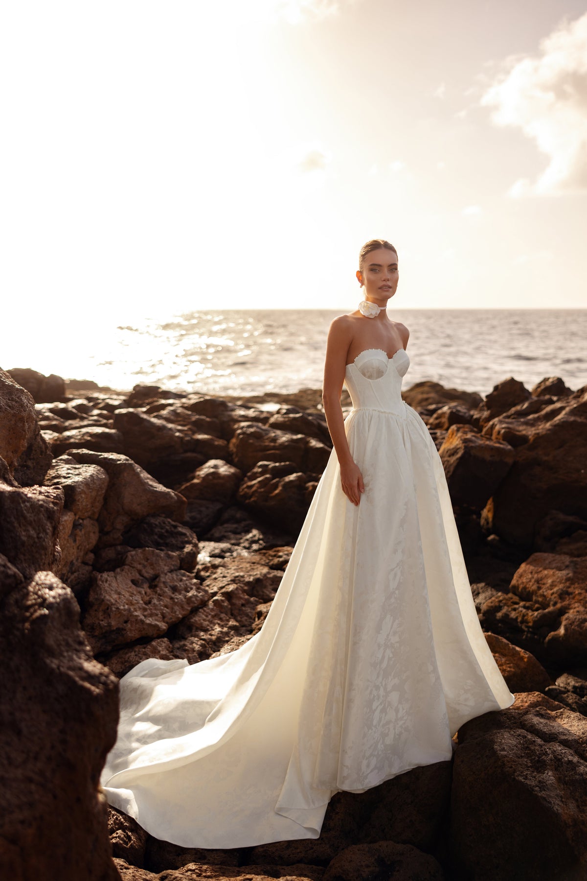 L’abito da sposa Cholly, con bustier strutturato e lungo strascico, fotografato al tramonto sul mare, esprime un’eleganza senza tempo.