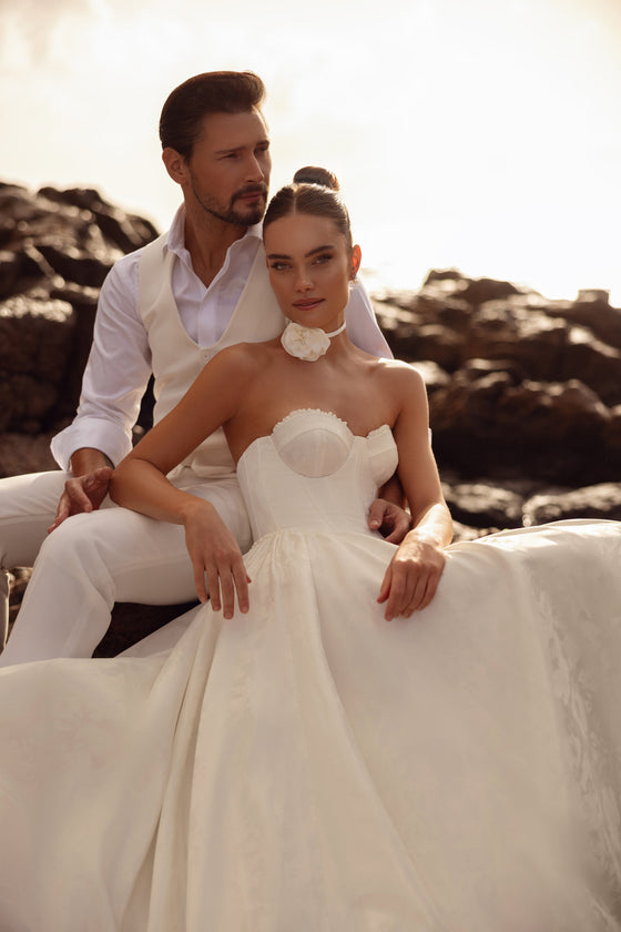 Abito da sposa Cholly dalla finitura satinata, con corpetto strutturato e gonna ampia, fotografato in un romantico servizio sul mare.