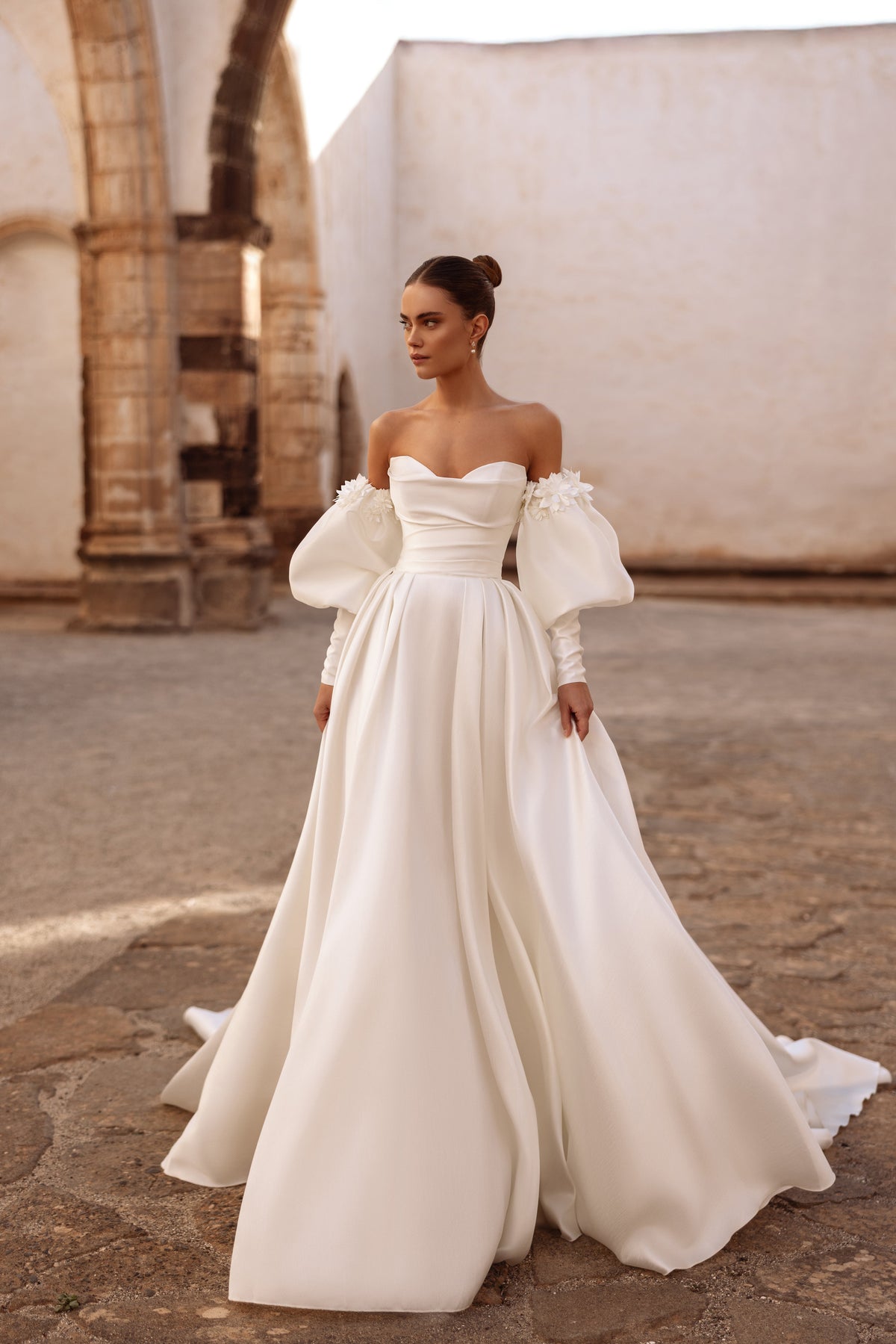Elegante abito da sposa Anika in raso, con spalle scoperte, maniche a sbuffo e applicazioni floreali, per un look romantico.