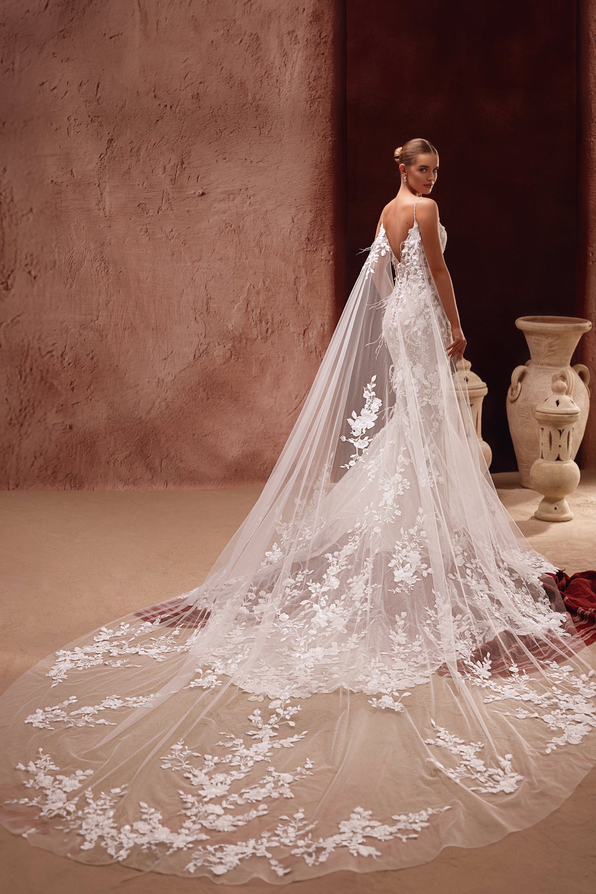 L’abito da sposa Aspasia, con scenografico strascico in pizzo e cappa ricamata a fiori, crea una figura indimenticabile.