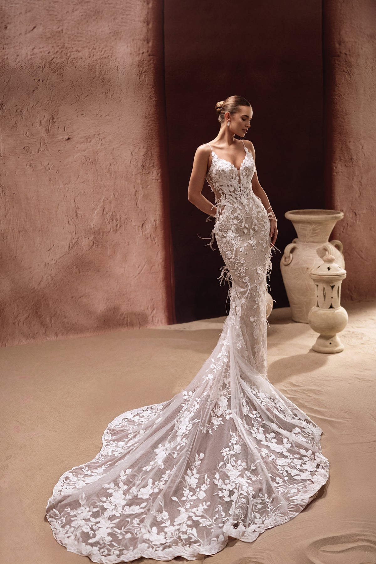L’abito da sposa a sirena Aspasia, con applicazioni di pizzo e dettagli di piume, valorizza le curve con un accento glamour couture.