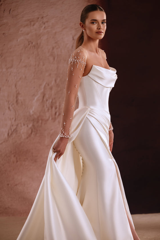 L’abito da sposa Aylin presenta maniche trasparenti impreziosite di perle e uno scollo drappeggiato in raso, per un look senza tempo.