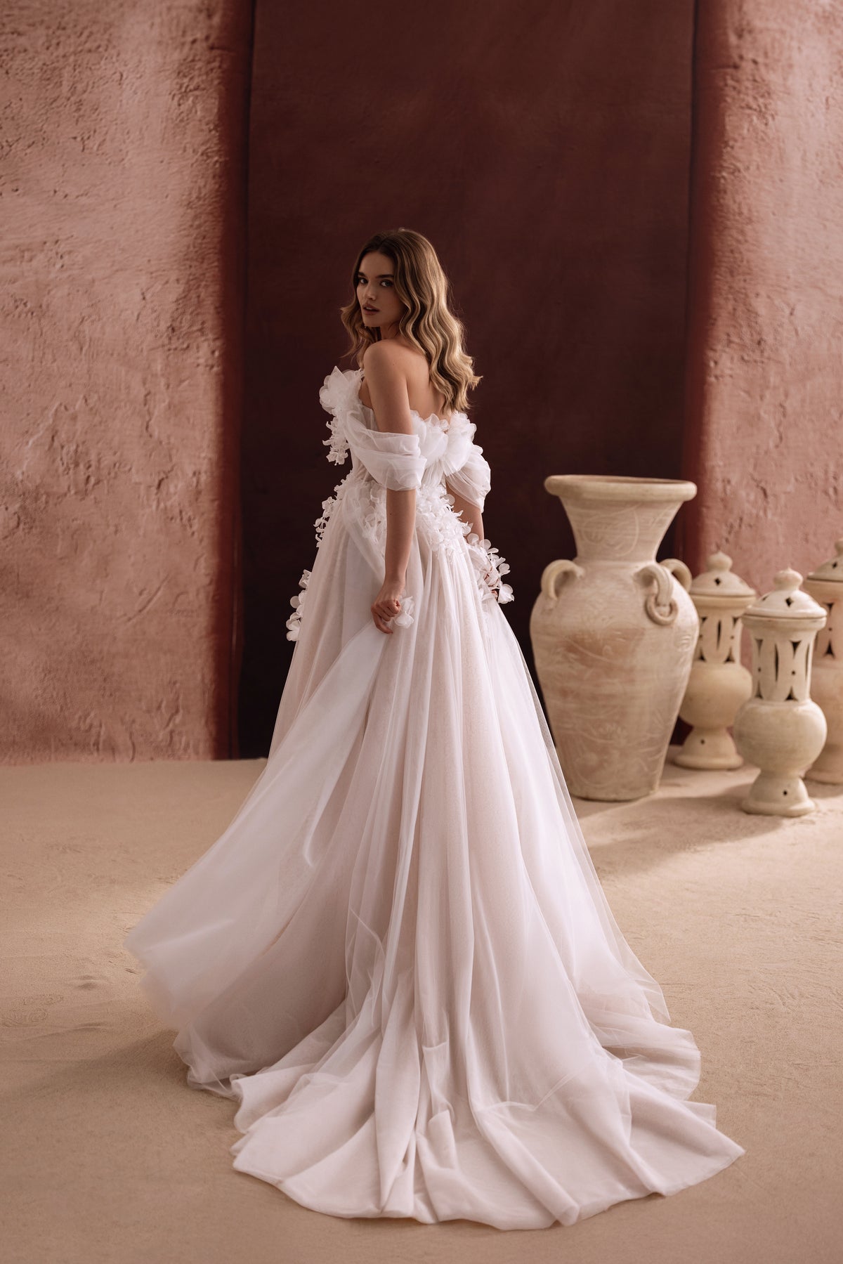 L’abito da sposa Bella, con maniche che lasciano scoperte le spalle e strascico fluido in tulle, disegna una figura romantica e sognante.