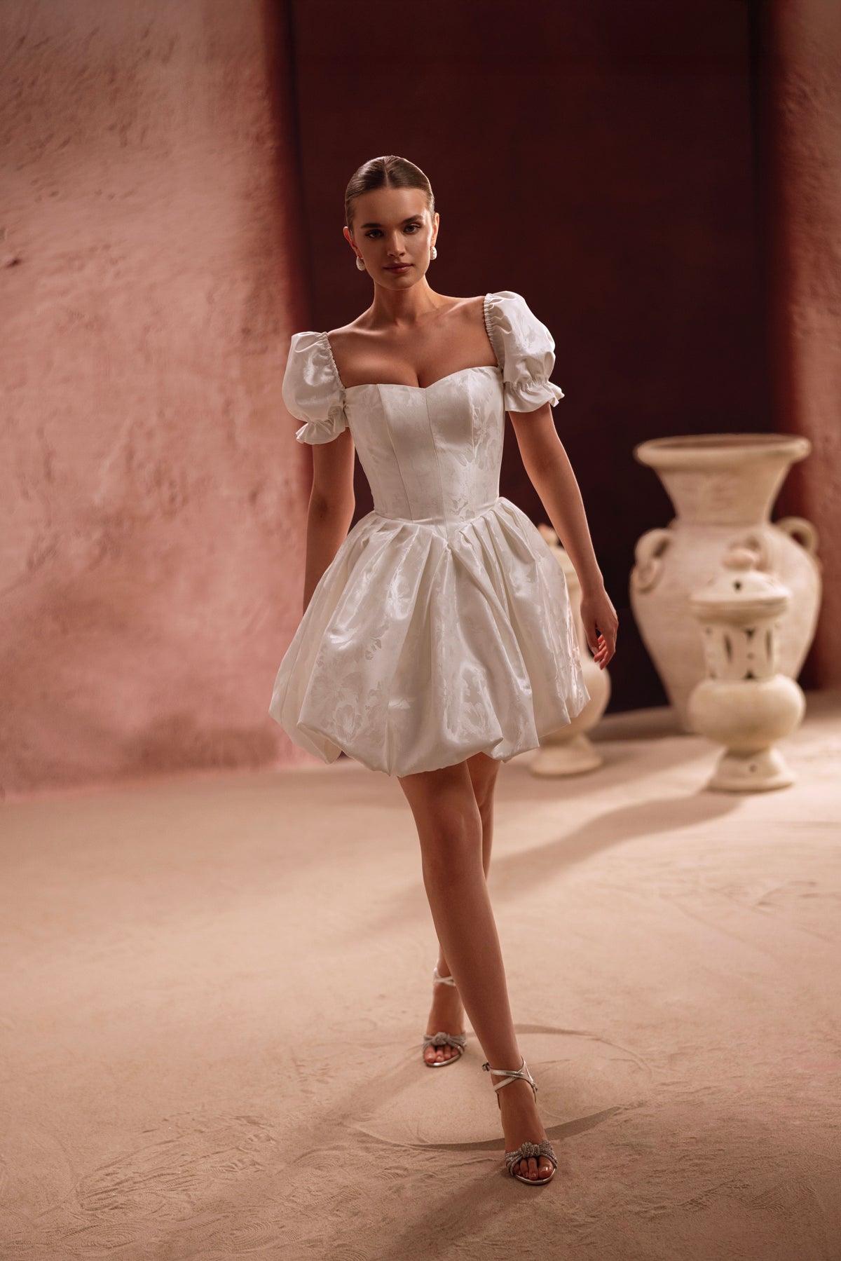 L’abito da sposa corto Daphne mostra un corsetto allacciato sulla schiena e maniche a sbuffo, unendo romanticismo e semplicità contemporanea.
