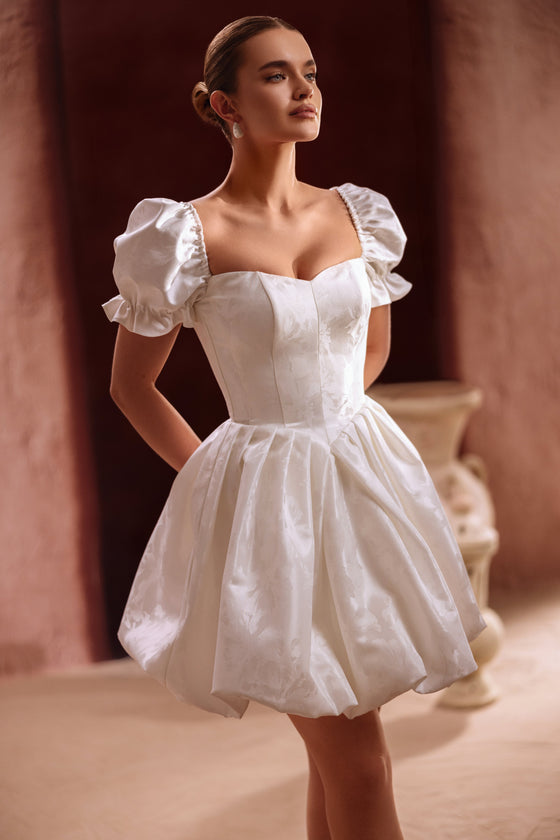L’abito da sposa corto Daphne presenta maniche a sbuffo e corpetto a corsetto, unendo fascino vintage e stile contemporaneo.