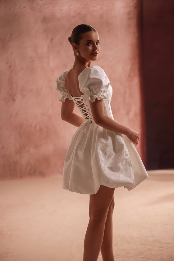 L’abito da sposa mini Daphne, con corsetto allacciato sulla schiena e maniche a sbuffo, sottolinea la vita e propone un giocoso look da principessa.