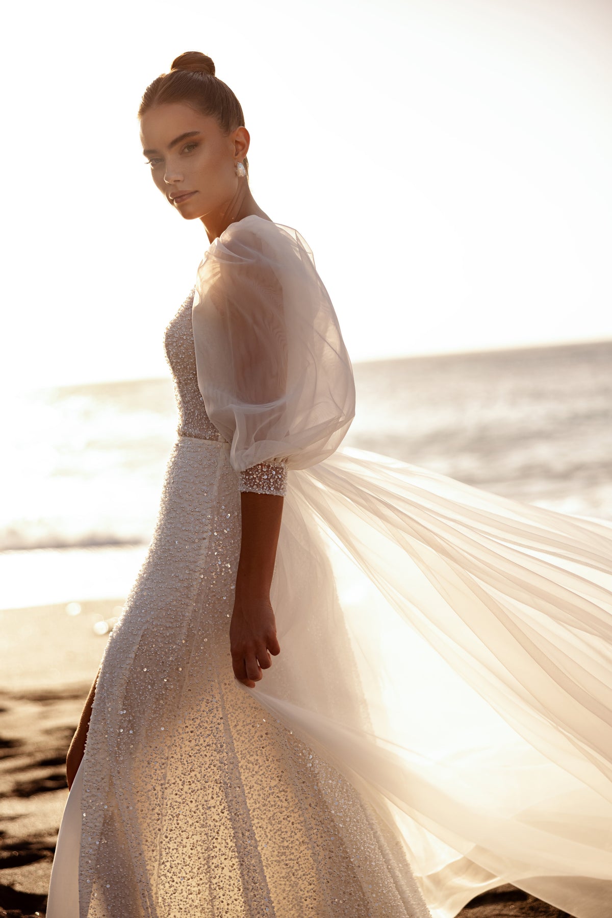 L’abito da sposa Davina, con tessuto scintillante e maniche voluminose, valorizza la femminilità con un accento glamour.