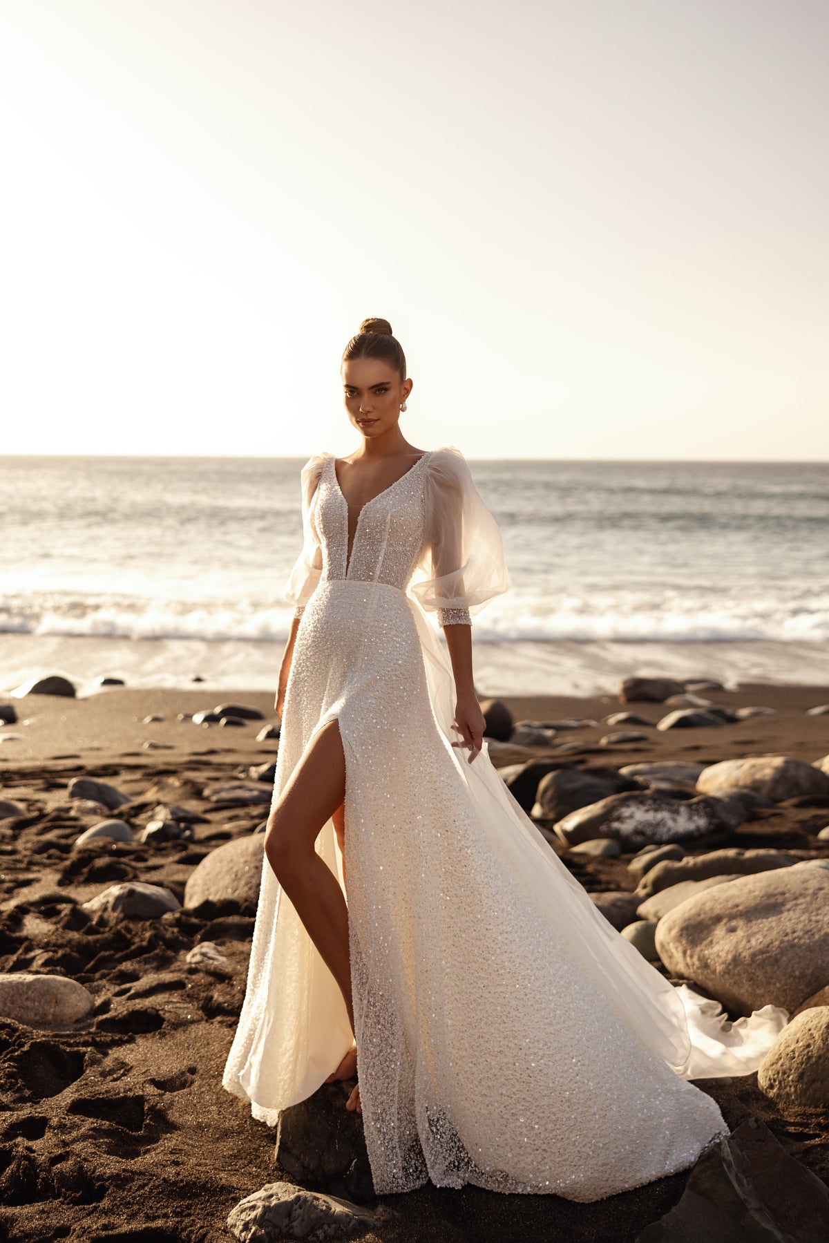 L’abito da sposa Davina presenta un profondo scollo a V e un tessuto scintillante, illuminato dal tramonto dorato sulla spiaggia.