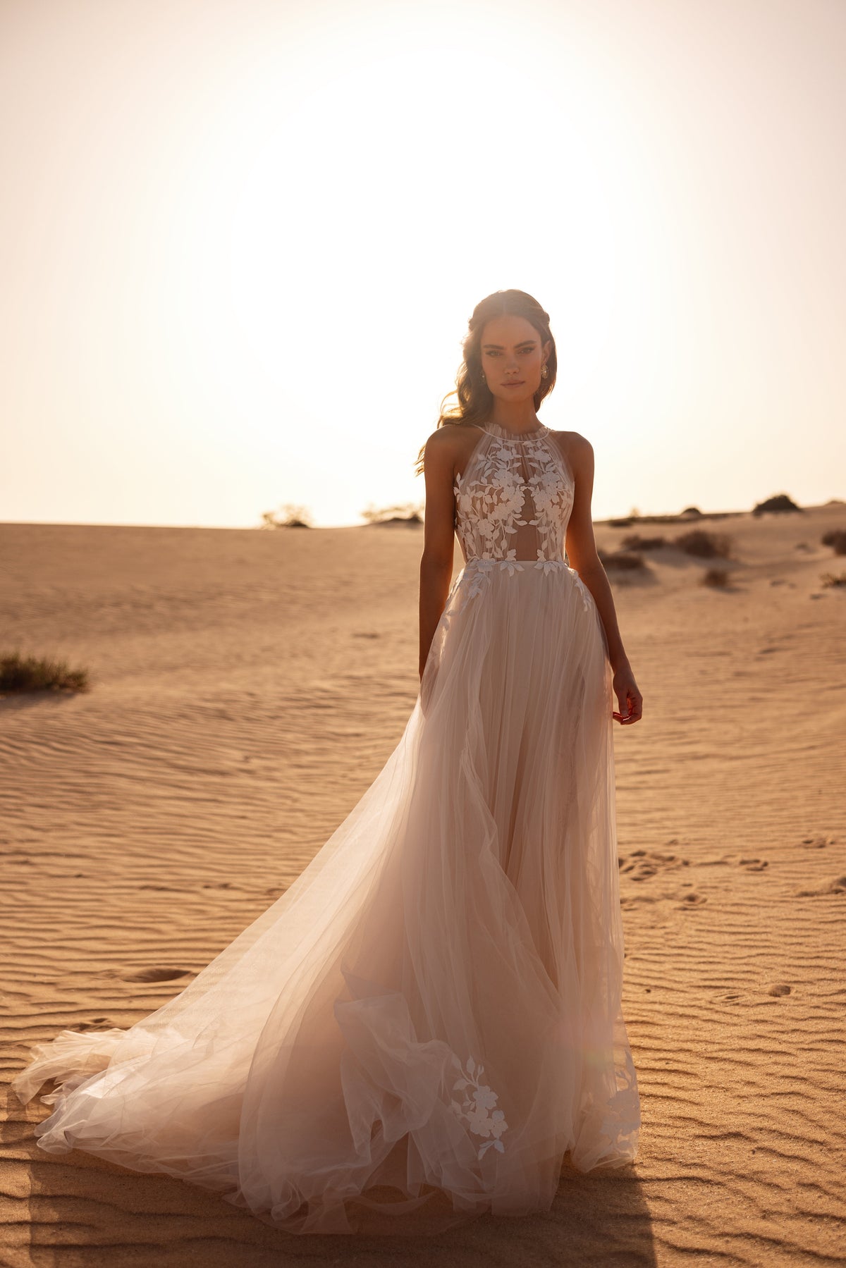 L’abito da sposa Ellie presenta un corpetto all’americana effetto illusion e una gonna fluida in tulle con applicazioni floreali, per un look romantico.