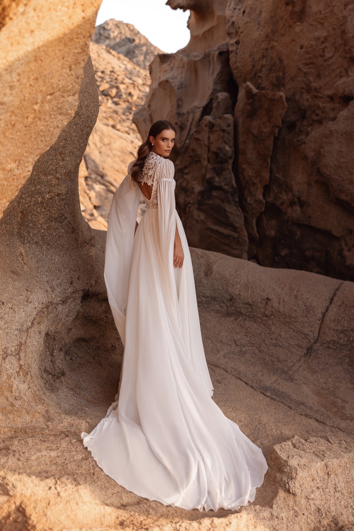 L’abito da sposa Etra presenta un profondo scollo a V, uno spacco scenografico e maniche a cappa in chiffon, per un look boho-chic.