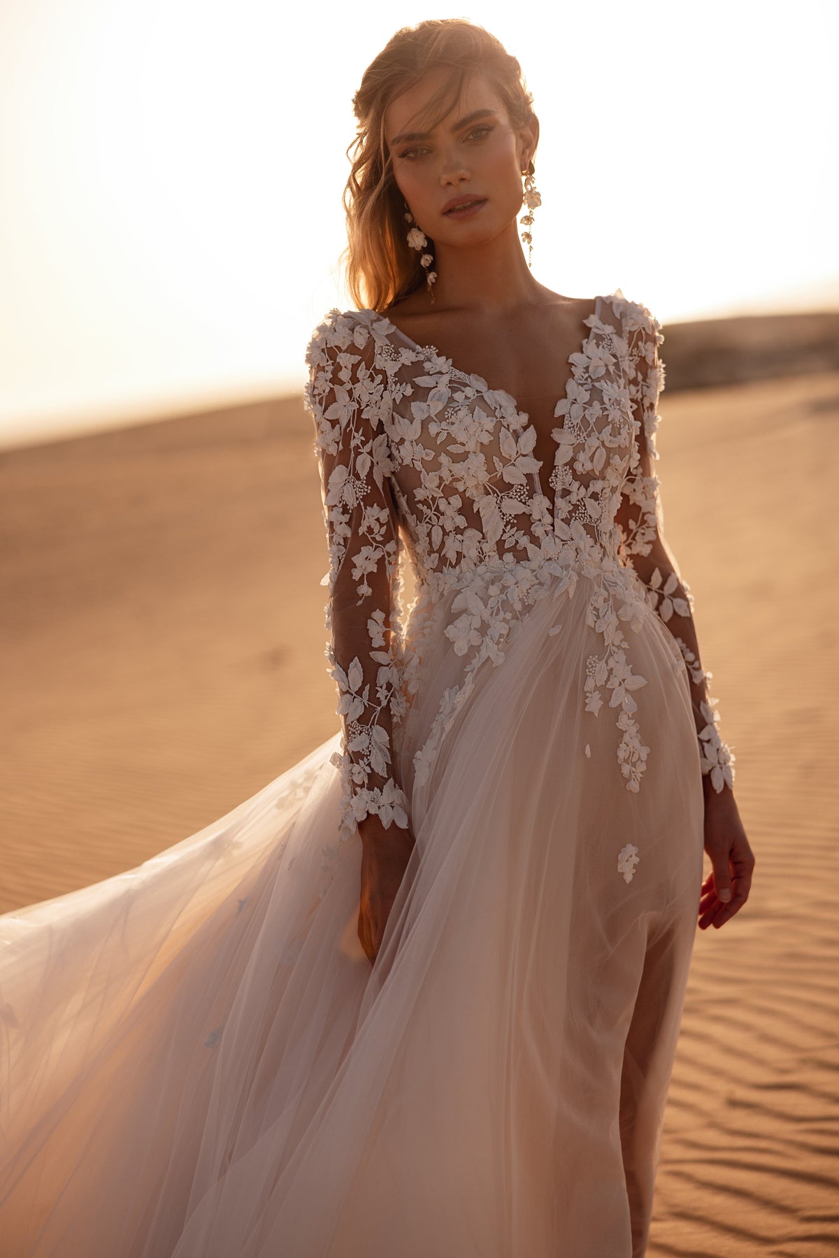 L’abito da sposa Farida presenta un profondo scollo a V, lunghe maniche in pizzo e delicate applicazioni floreali 3D, per un look romantico.