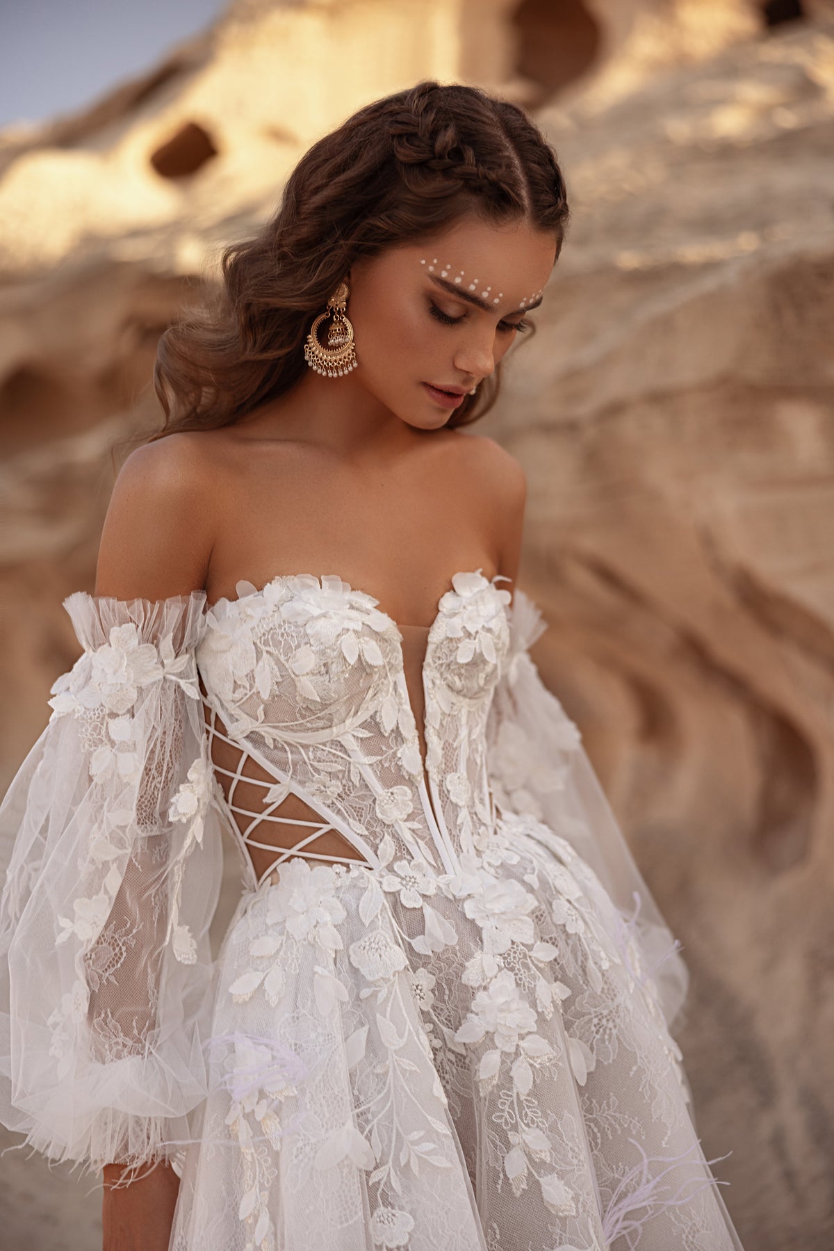 L’abito da sposa Huda presenta maniche che lasciano scoperte le spalle e un corsetto strutturato in pizzo, per un look boho glamour contemporaneo.