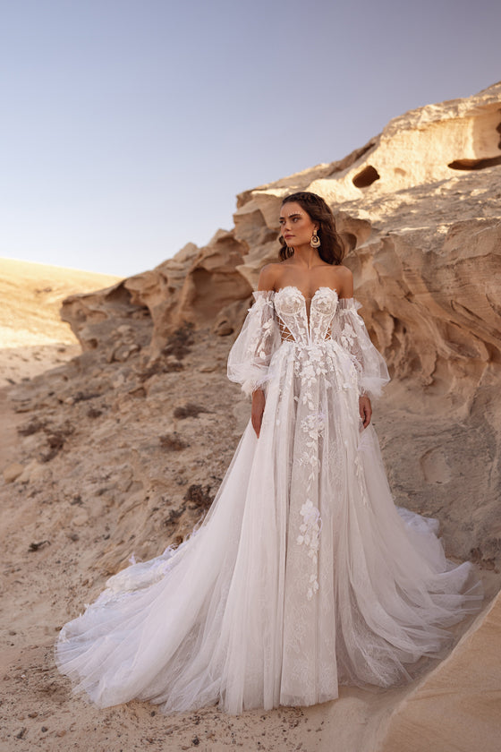 L’abito da sposa Huda presenta un corsetto a spalle scoperte con applicazioni floreali 3D e allacciatura laterale, per un look boho contemporaneo.