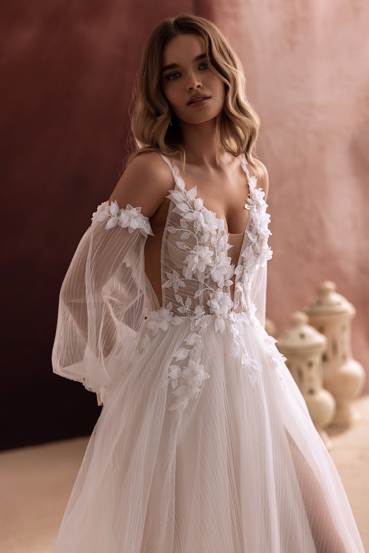 L’abito da sposa Lilibeth, con scollo profondo e romantiche maniche a sbuffo in tulle annodate con fiocchi, sottolinea l’eleganza femminile.