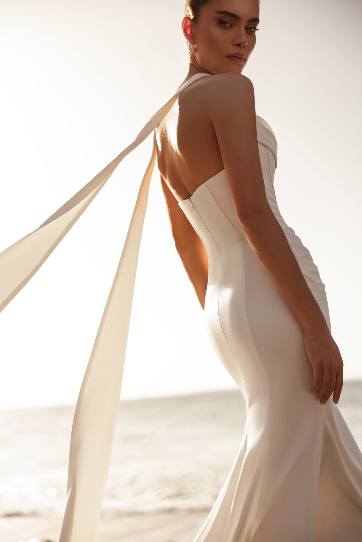 Il lungo strascico e i nastri fluidi dell’abito da sposa Luna creano un look contemporaneo d’effetto in riva all’oceano.
