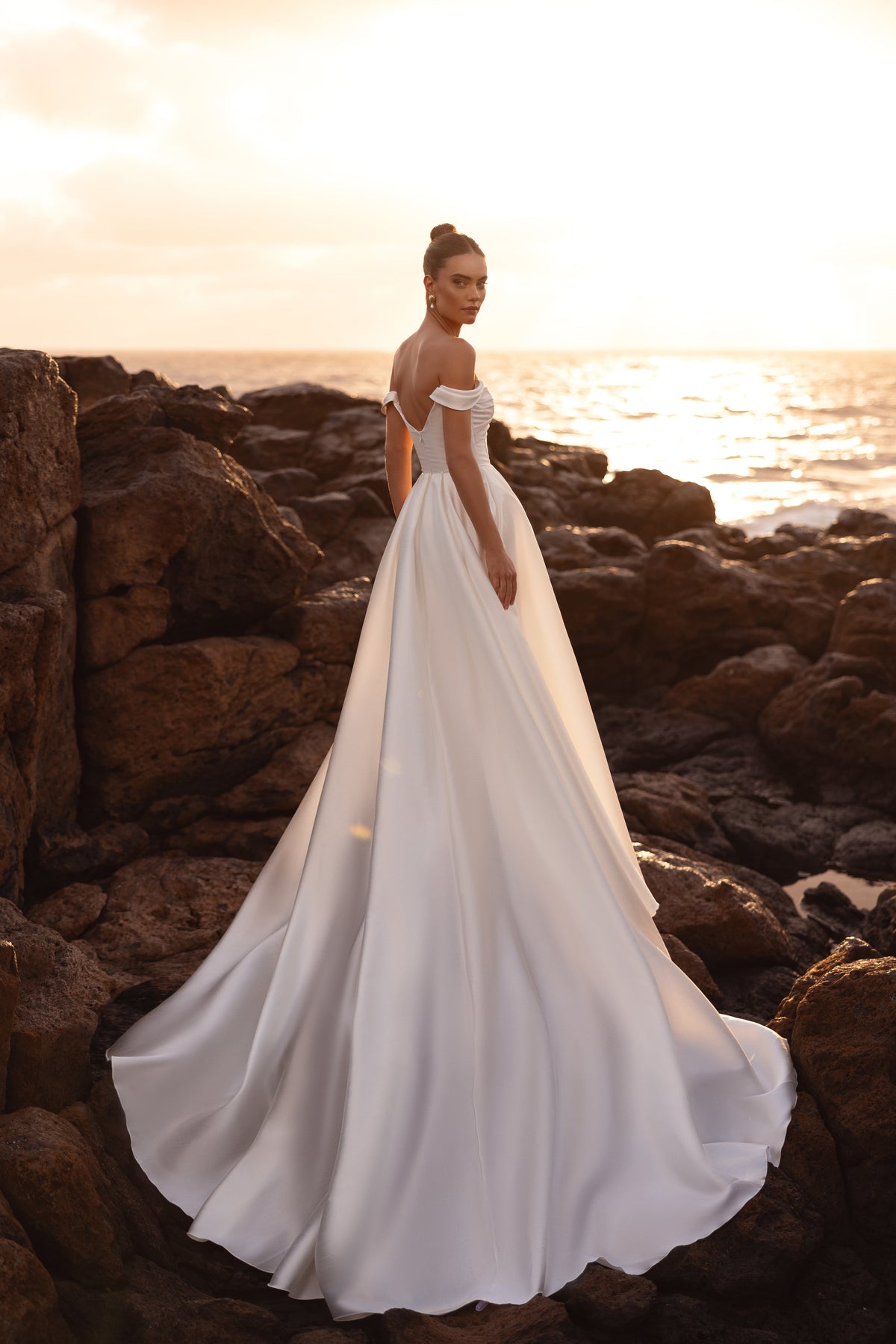 L’abito da sposa Mellisandre, con spalle scoperte e strascico scenografico illuminato dal tramonto, si presta a matrimoni sulla spiaggia o all’aperto.