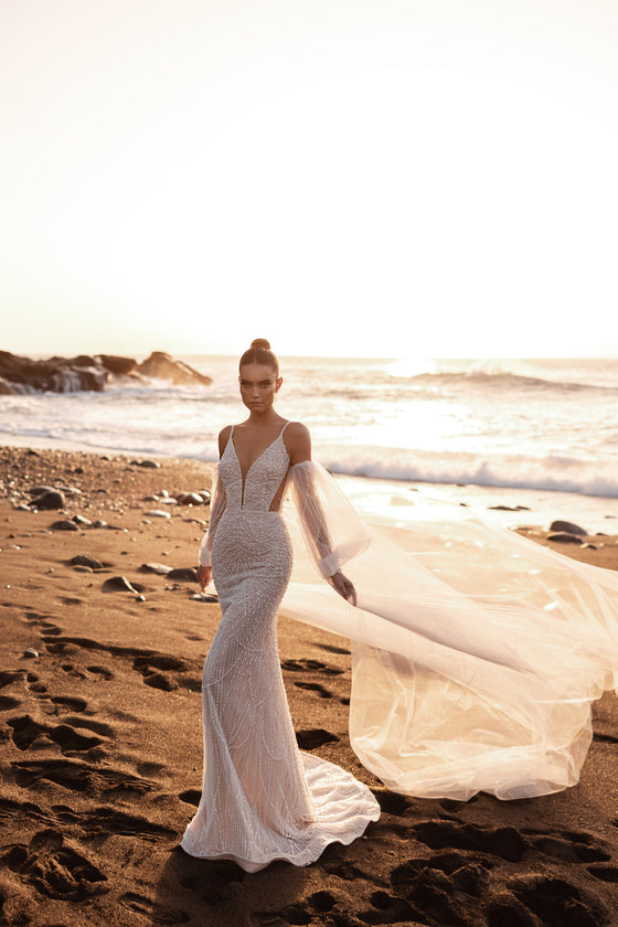 Abito da sposa a sirena aderente, con profondo scollo a V e velo fluido di lunghezza cappella, sulla spiaggia.