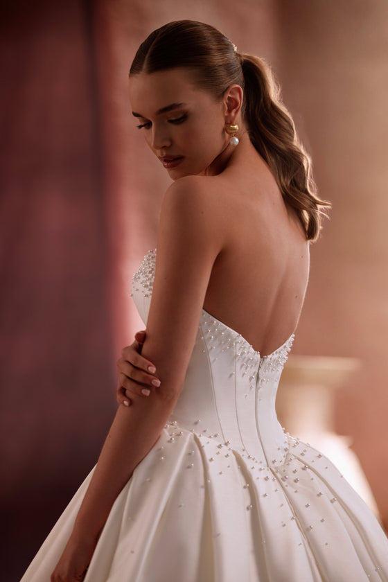 L’abito da sposa Noor in raso, con gonna ampia e strascico scenografico, mostra una luminosità preziosa e un disegno d’ispirazione couture.