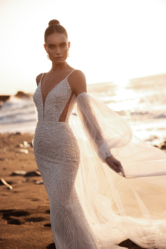 L’abito da sposa Rebecca a sirena, con spalline sottili e ricami di perle, risplende al tramonto, per un look sulla spiaggia senza tempo.