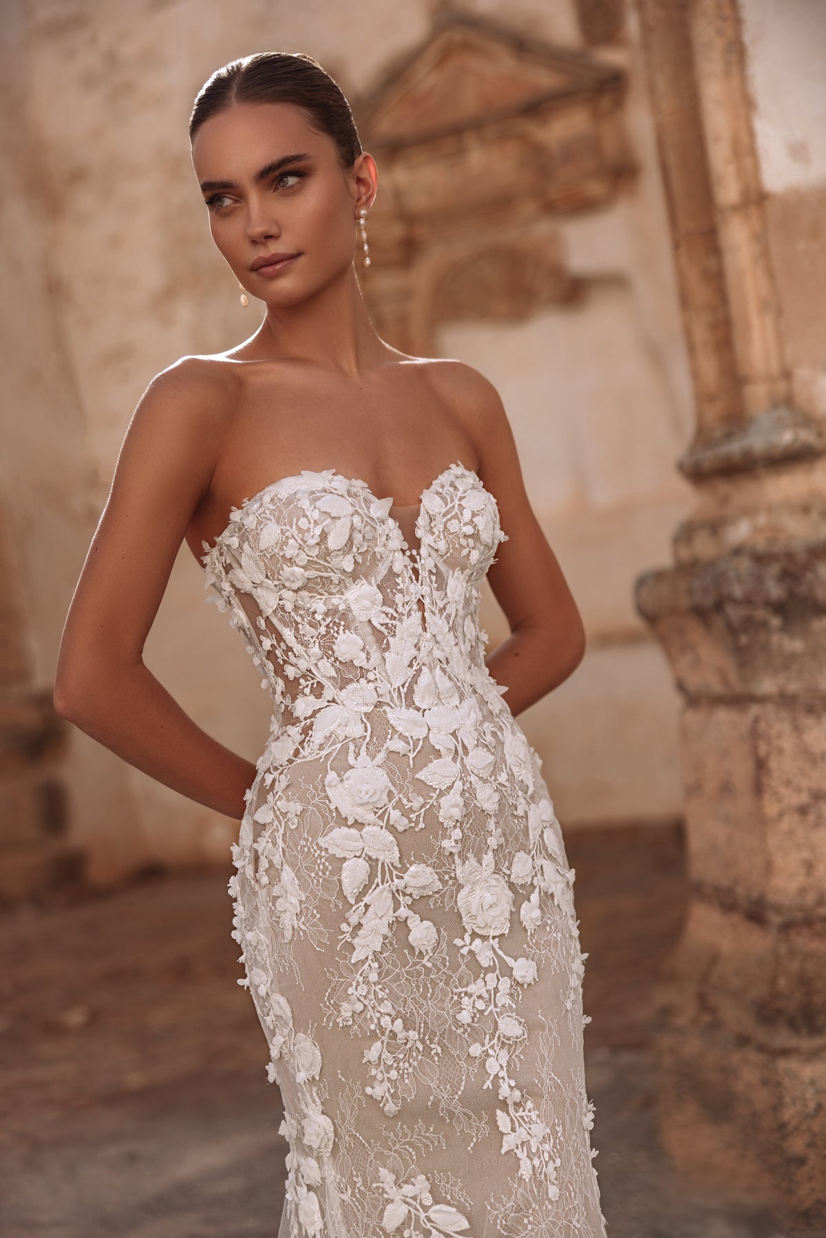 L’abito da sposa Sarifa in pizzo, con strascico scenografico e dettagli floreali, esprime lusso e romanticismo contemporaneo.
