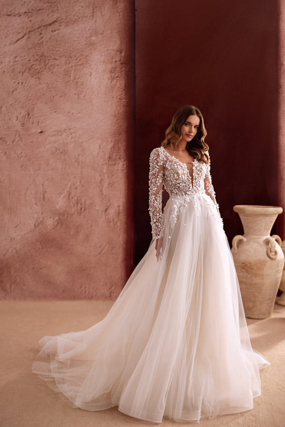 L’abito da sposa Shanin, con lunghe maniche in pizzo, scollo profondo e gonna fluida in tulle, esprime romanticismo disinvolto ed eleganza contemporanea.