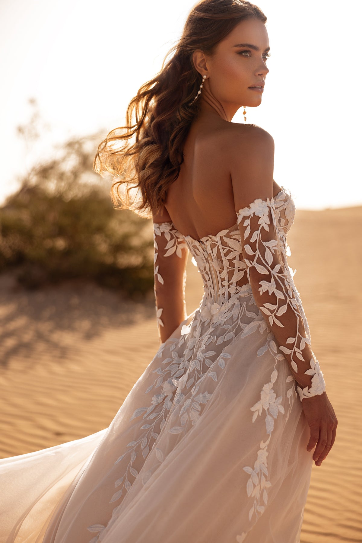 L’abito da sposa Tamara, con maniche che lasciano scoperte le spalle e applicazioni di pizzo floreale, crea un look sensuale e femminile.