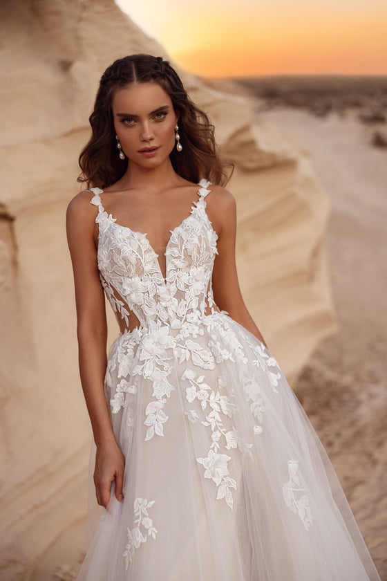L’abito da sposa Zafira, con gonna in tulle e dettagli in pizzo floreale, accanto a uno sposo in completo beige, crea un’immagine sognante.